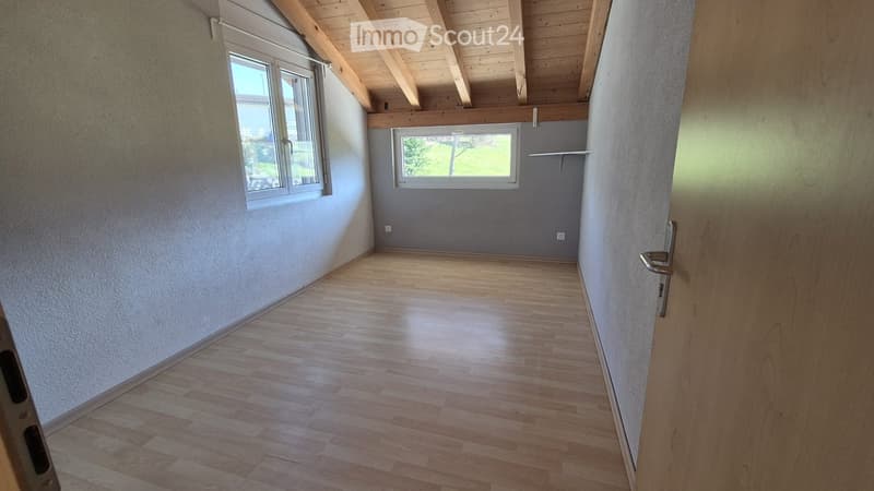 Empty room with wooden flooring, exposed wooden beams on the ceiling, and large windows allowing natural light to enter.