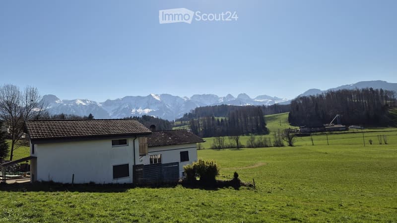 A single-story white house with a tiled roof, situated in a green meadow with snow-capped mountains in the background.