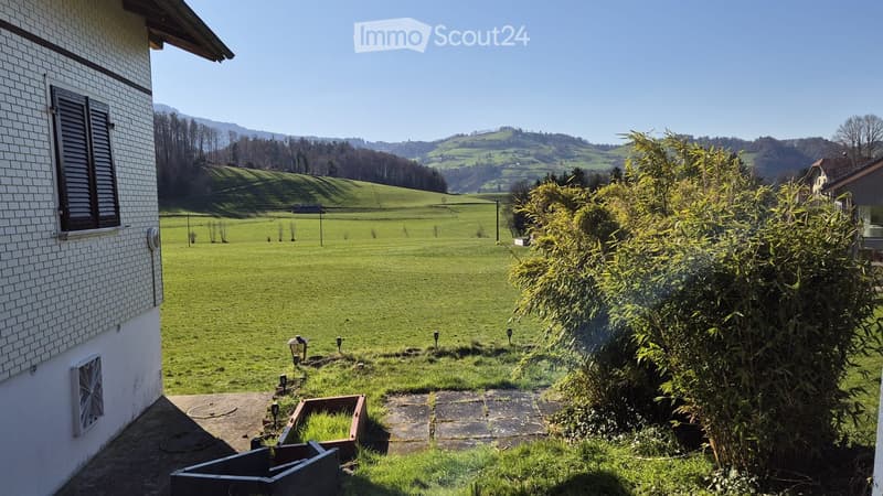 A peaceful garden area with a well-maintained lawn, surrounded by trees and hills in the background. The scene includes a paved pathway and a small garden bed near the house.