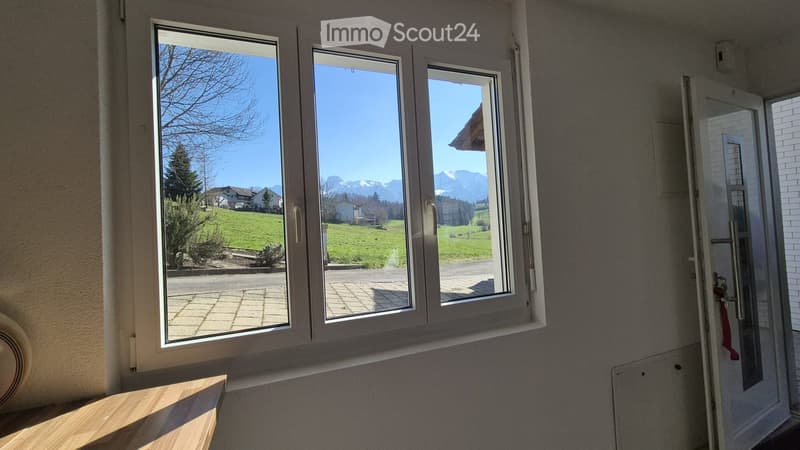 Empty room with large windows offering a scenic view of a green landscape and mountains. The room features a wooden floor and a door leading outside.