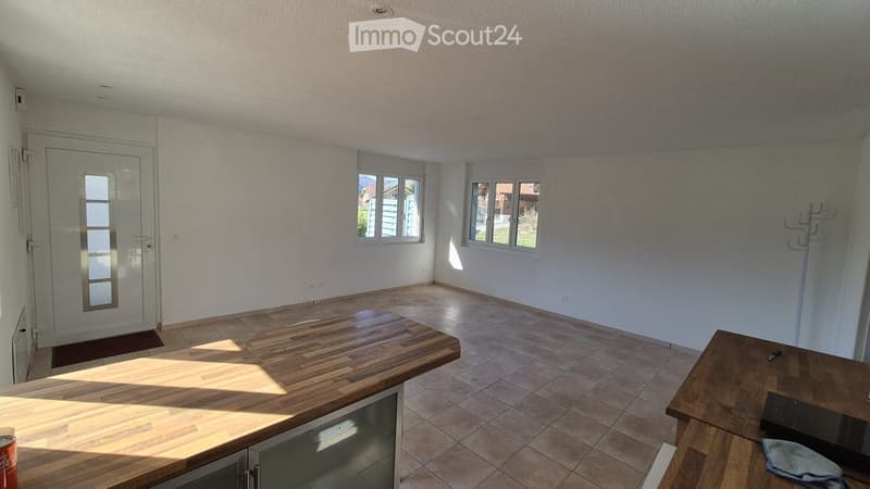 A bright, unfurnished room with a wooden table, a chair, a door, and windows letting in natural light.