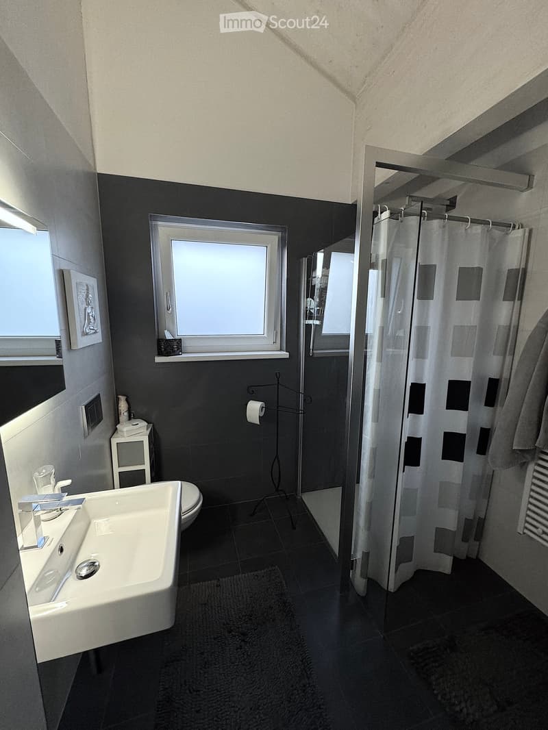 Modern bathroom, gray tiles, white countertop, undermount sink, shower enclosure, toilet, window, decorative items, and rugs.