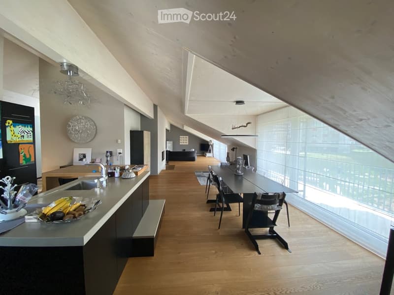 Open-plan kitchen, dining, and living area with wooden floors and vaulted ceiling. Large glass windows and balconies with views. Modern furnishings and appliances.