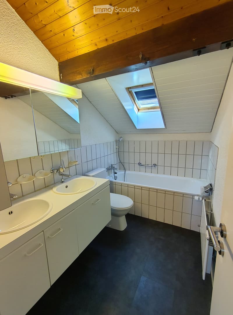 Modern bathroom with a double sink, toilet, bathtub, and large mirror. The room features wooden slanted ceilings and skylights for natural light.