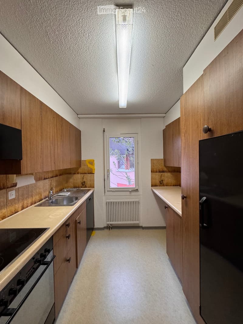 Compact kitchen with wooden cabinets, a sink, oven, and refrigerator. Features a window with a view of a neighboring building.
