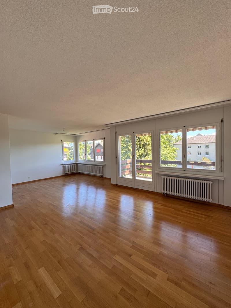 Empty room with wooden flooring, large windows, radiators, and sliding glass doors leading to a balcony.