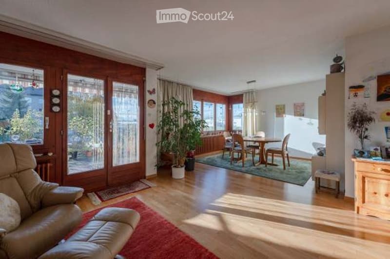 Bright living and dining area with large windows, wooden flooring, a leather sofa, ottoman, dining table with chairs, potted plants, and a cabinet.