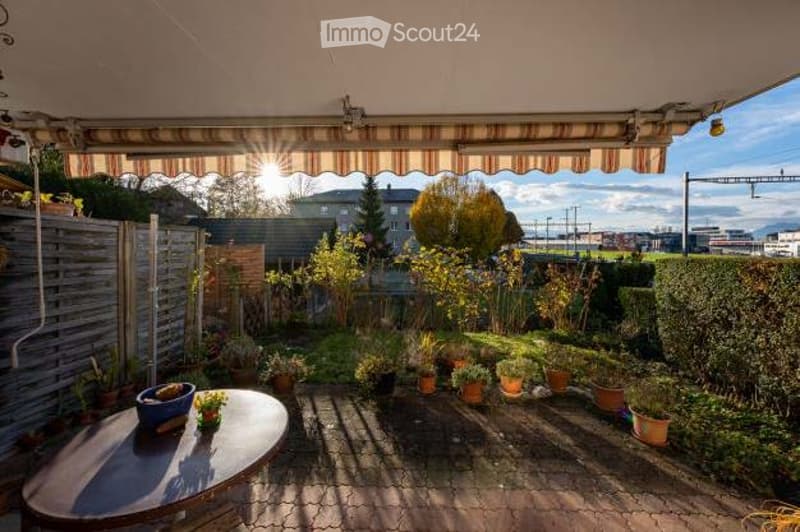 A sunny patio with a round wooden table, potted plants, and a wooden fence, surrounded by greenery and a view of the city.