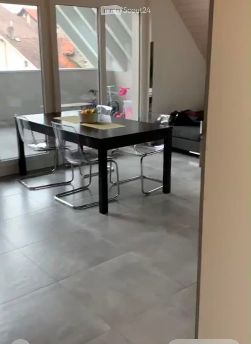 Modern dining area with a black dining table, four chairs, and a bowl of fruit on the table. Large windows provide natural light and a view of the outdoor area.