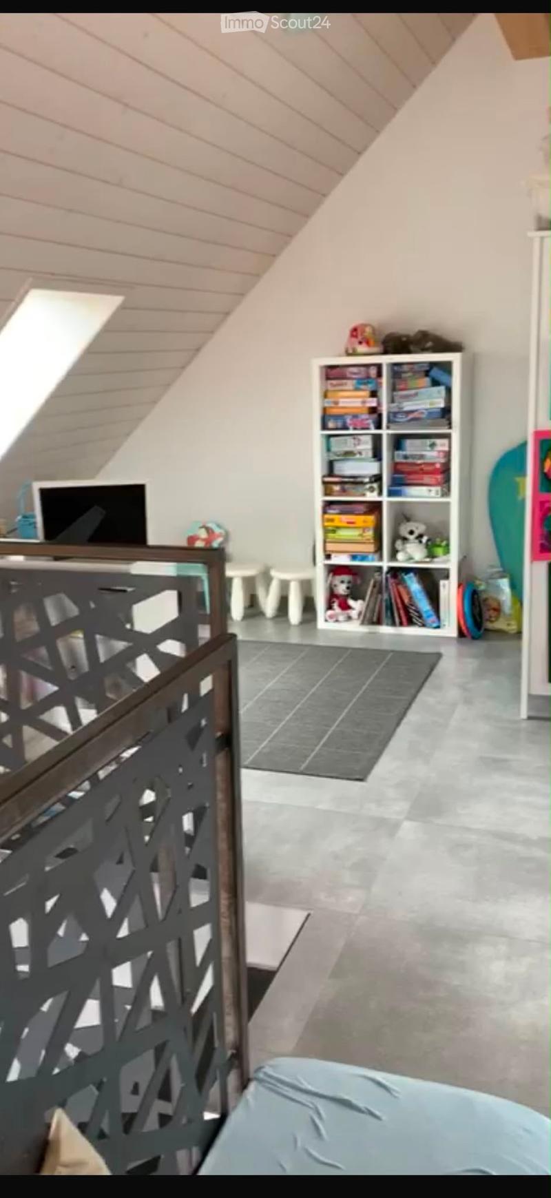 A furnished room with a slanted ceiling, a screen, a small table, a shelf filled with books and toys, and a stool.
