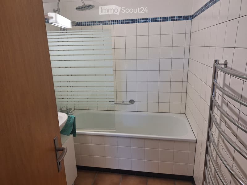 Modern bathroom with white tiled walls, a bathtub with a glass shower enclosure, a towel rack, and a sink with a green towel.