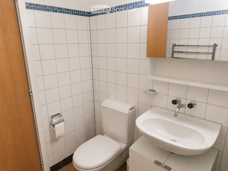 Modern bathroom with white tiles, a toilet, a sink with a chrome faucet, a wooden cabinet, a towel rack, and a shelf above the sink.