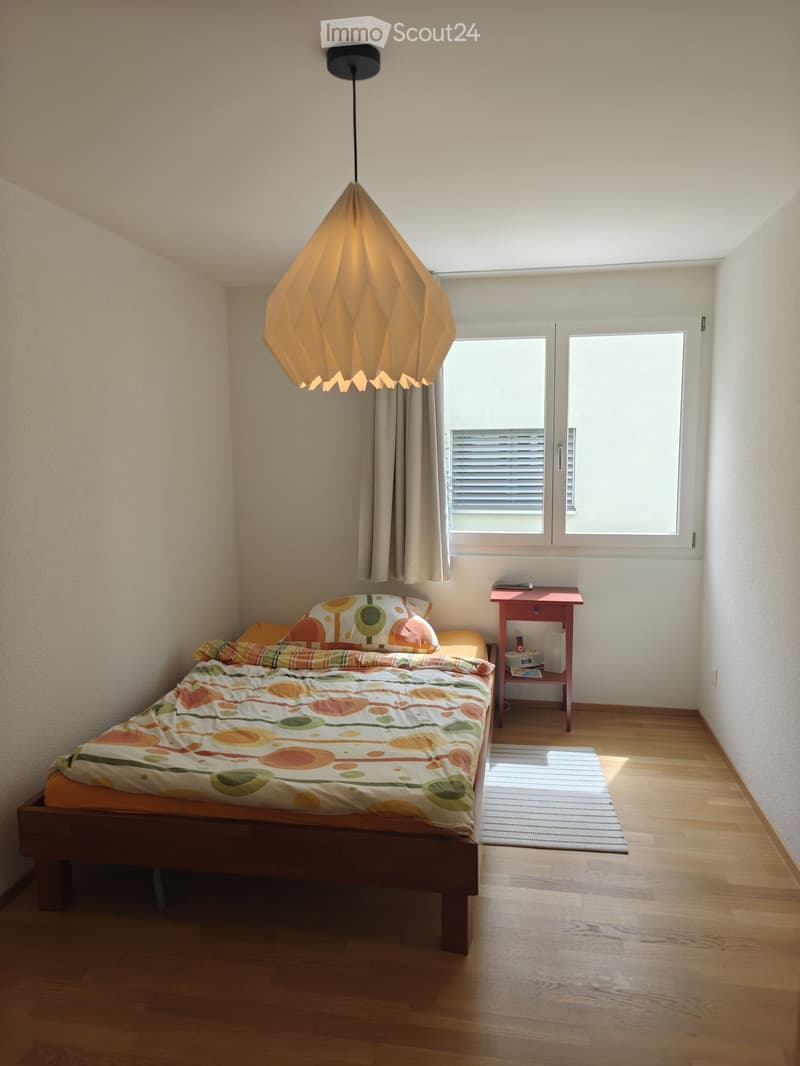 A cozy bedroom with a wooden bed, colorful bedding, a nightstand, and a hanging geometric lamp. The room features wooden flooring and large windows with blinds.