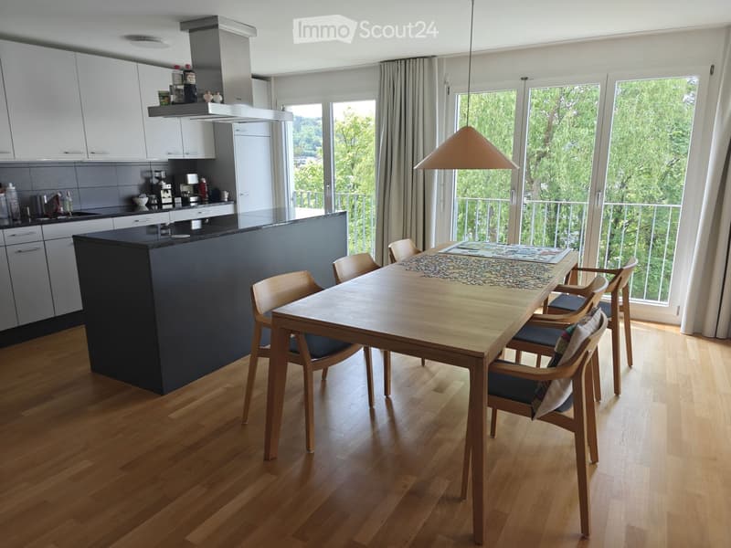 Open-plan kitchen and dining area with wooden flooring, modern kitchen cabinets, a central island, and a dining table with chairs. Large windows offer a view of greenery.
