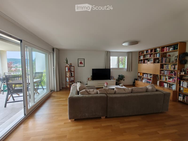 Modern living room with wooden flooring, large sliding glass doors leading to a balcony, a grey sectional sofa, coffee table, bookshelf, and television.