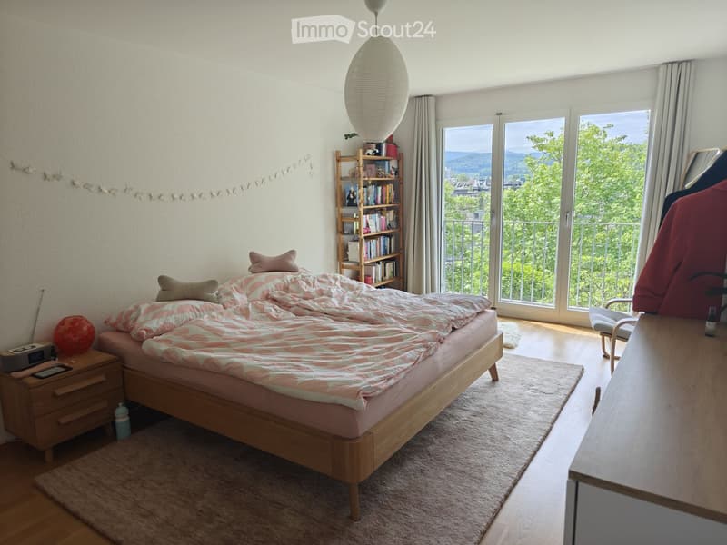 Cozy bedroom with a wooden bed, nightstand, bookshelf, and large windows offering a scenic view. Includes a chair, table, and decorative lamp.