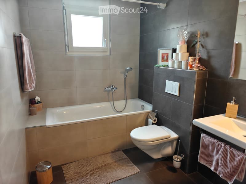 Modern bathroom with a bathtub, toilet, sink, and various accessories including towels, a mirror, and a candle. The space features a window and a dark grey accent wall.