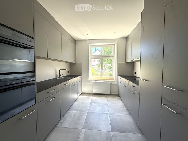 Modern kitchen with grey cabinets, black countertops, and a large window letting in natural light. Features a sink, faucet, and built-in oven.