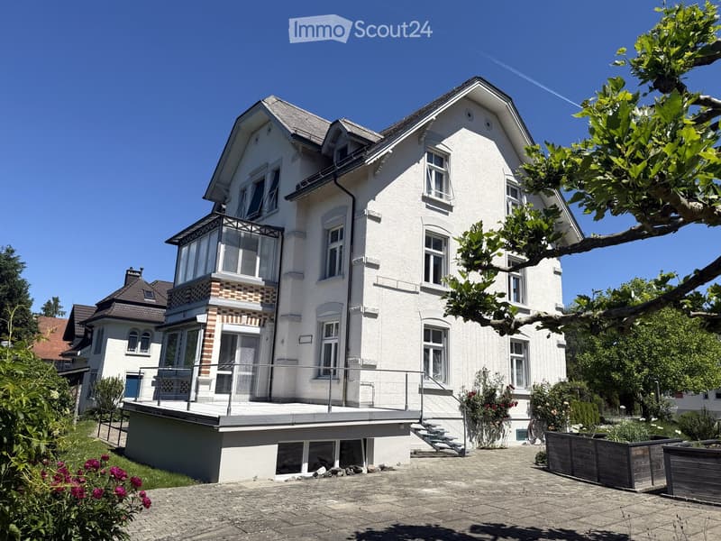 A two-story white house with multiple windows, a balcony, and a well-maintained garden area.