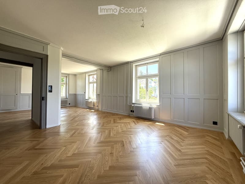 Empty room with wooden herringbone flooring, white walls, large windows, radiators, and a door leading to another room.