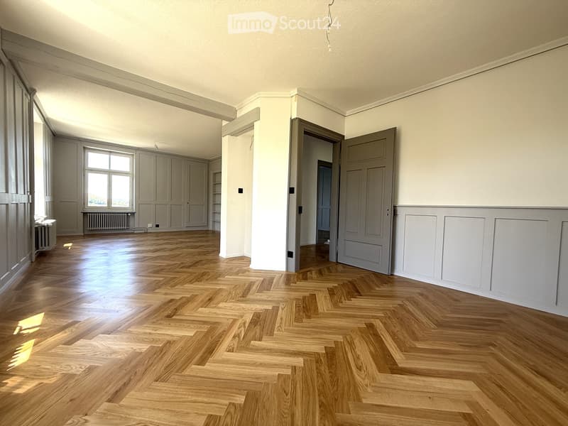 Empty room with wooden herringbone flooring, white walls, large window, and several doors leading to other spaces.