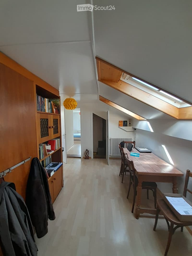 A narrow hallway with wooden flooring leads to a small, furnished room with a wooden table, chairs, a laptop, and a bookshelf. The room has slanted ceilings with skylights and a radiator.