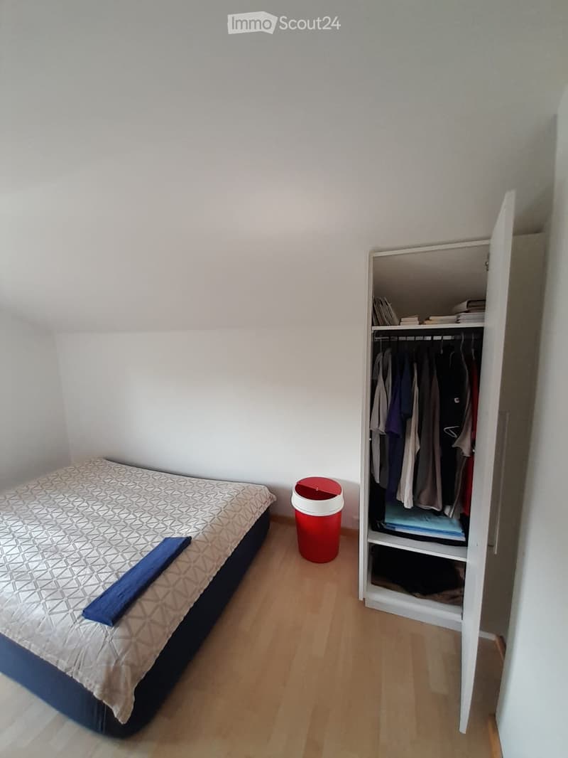 A small bedroom with a single bed, a wardrobe with hanging clothes, and a red trash bin on the floor.