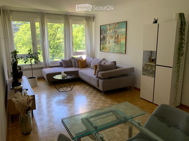 Modern living room with a grey sectional sofa, glass coffee table, glass side table, wooden sideboard, and a potted plant near the window.