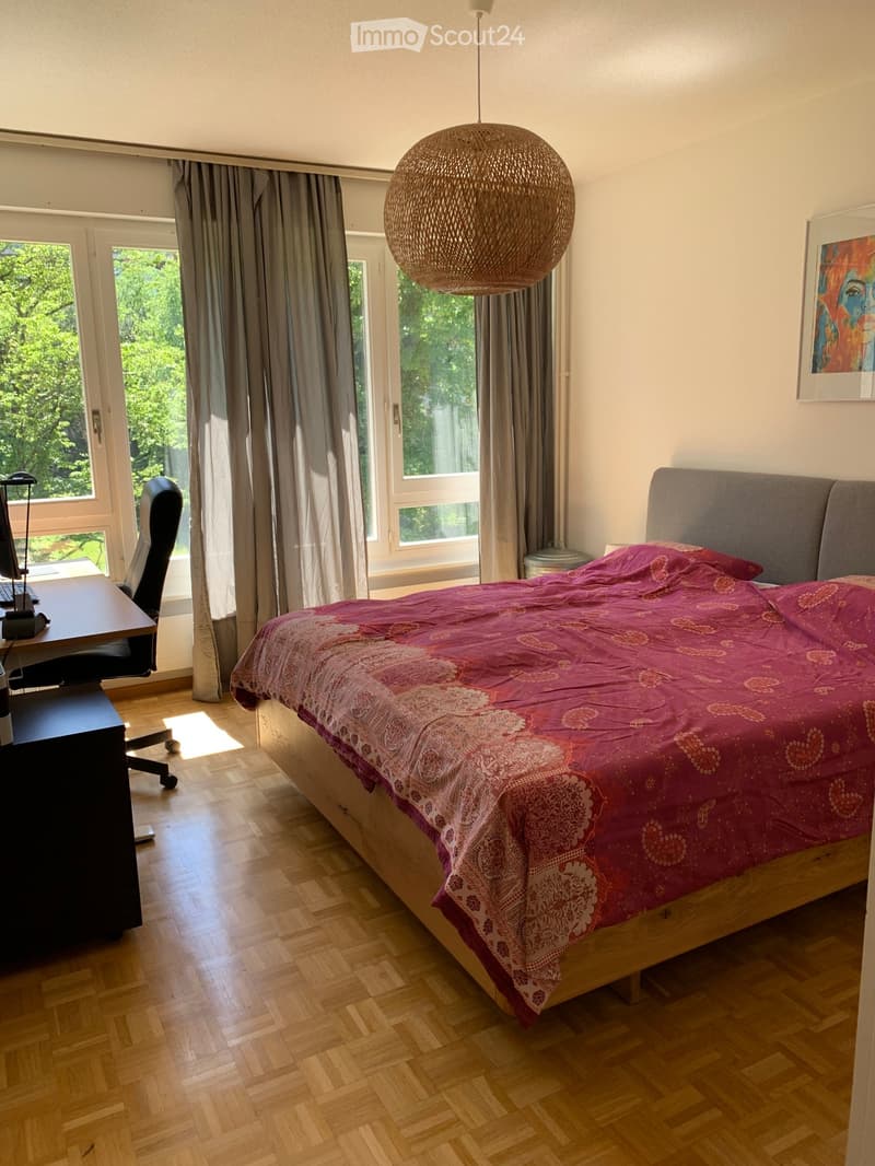 Bright bedroom with wooden flooring, large windows with grey curtains, a bed with a pink patterned duvet, a desk with a chair, and a hanging spherical lamp.