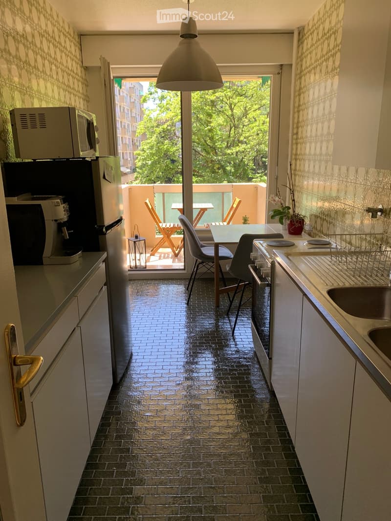 Modern kitchen with dark tiled floor, white cabinets, stainless steel appliances, and a small dining area leading to a balcony with outdoor furniture.