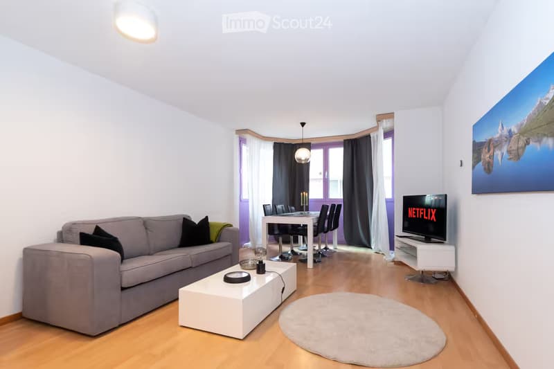 Modern living and dining area with light wooden flooring, white walls, grey sofa, coffee table, dining table with chairs, television, and artwork on the wall.