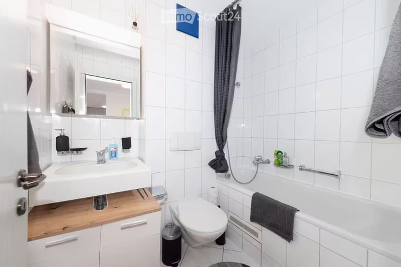 Modern bathroom with white tiles, a sink with a wooden countertop, a mirror, a toilet, a bathtub with a towel, and a grey curtain.
