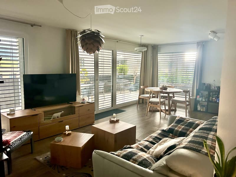 Bright living room with large windows and sliding doors leading to a balcony. Features a sofa, coffee table, TV, dining area with chairs and table, and decorative lamps.