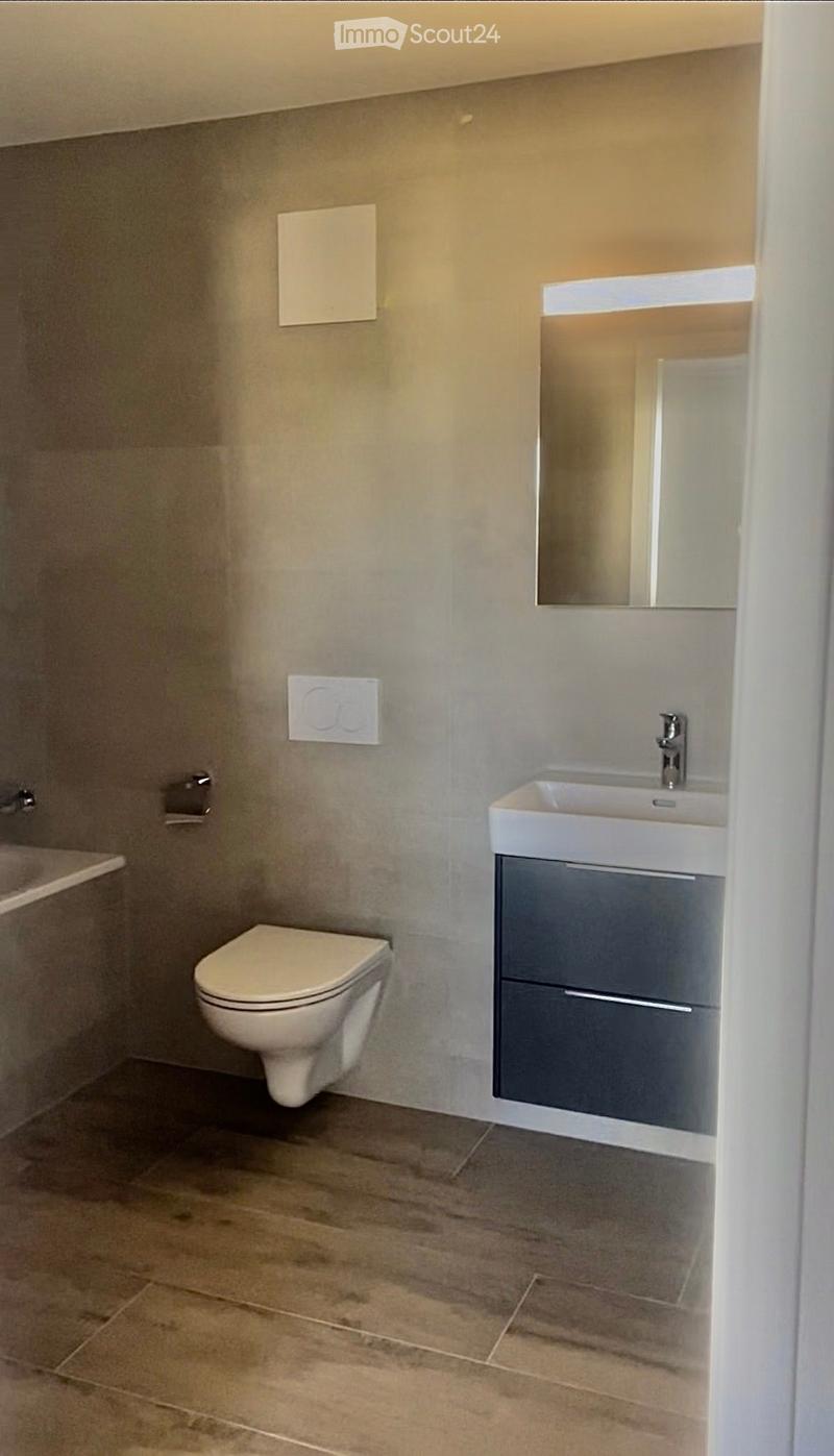Modern bathroom with a toilet, sink, mirror, and cabinet. The walls are light grey, and the floor is tiled.