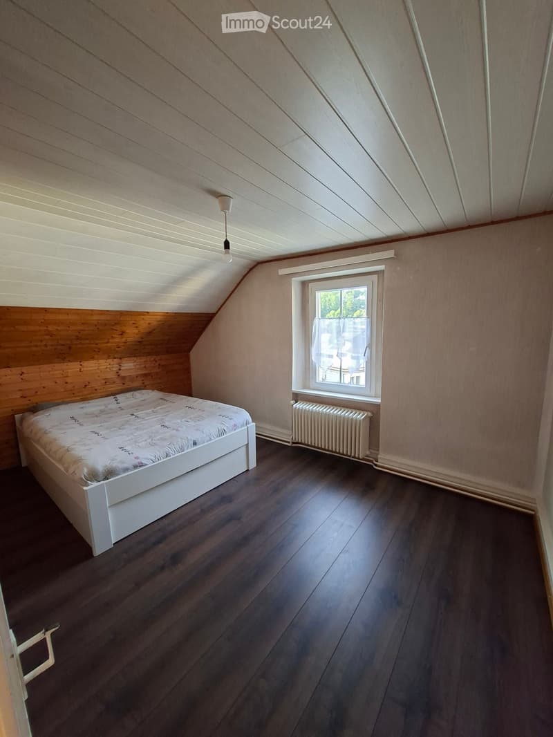Bedroom with wooden floor, slanted ceiling, white bed with patterned bedding, radiator under window, and a window with white frame and curtain.