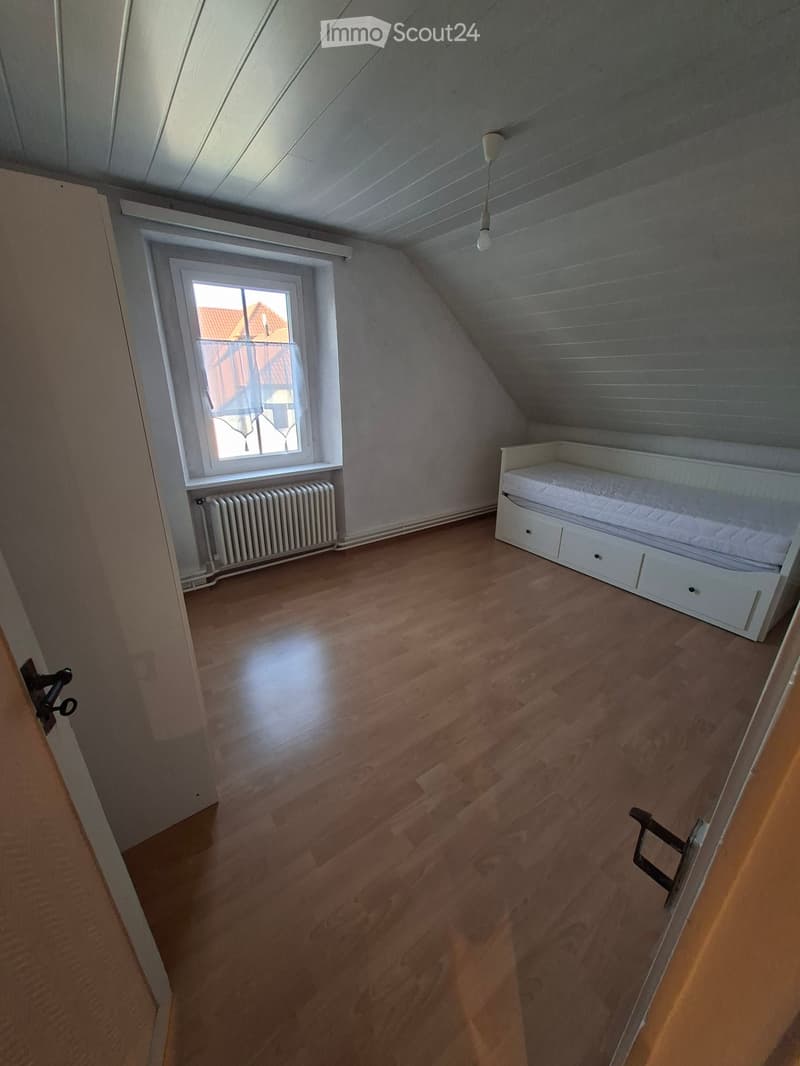 A small attic bedroom with wooden flooring, a single bed with drawers, a window with a radiator below, and a slanted ceiling with exposed beams.