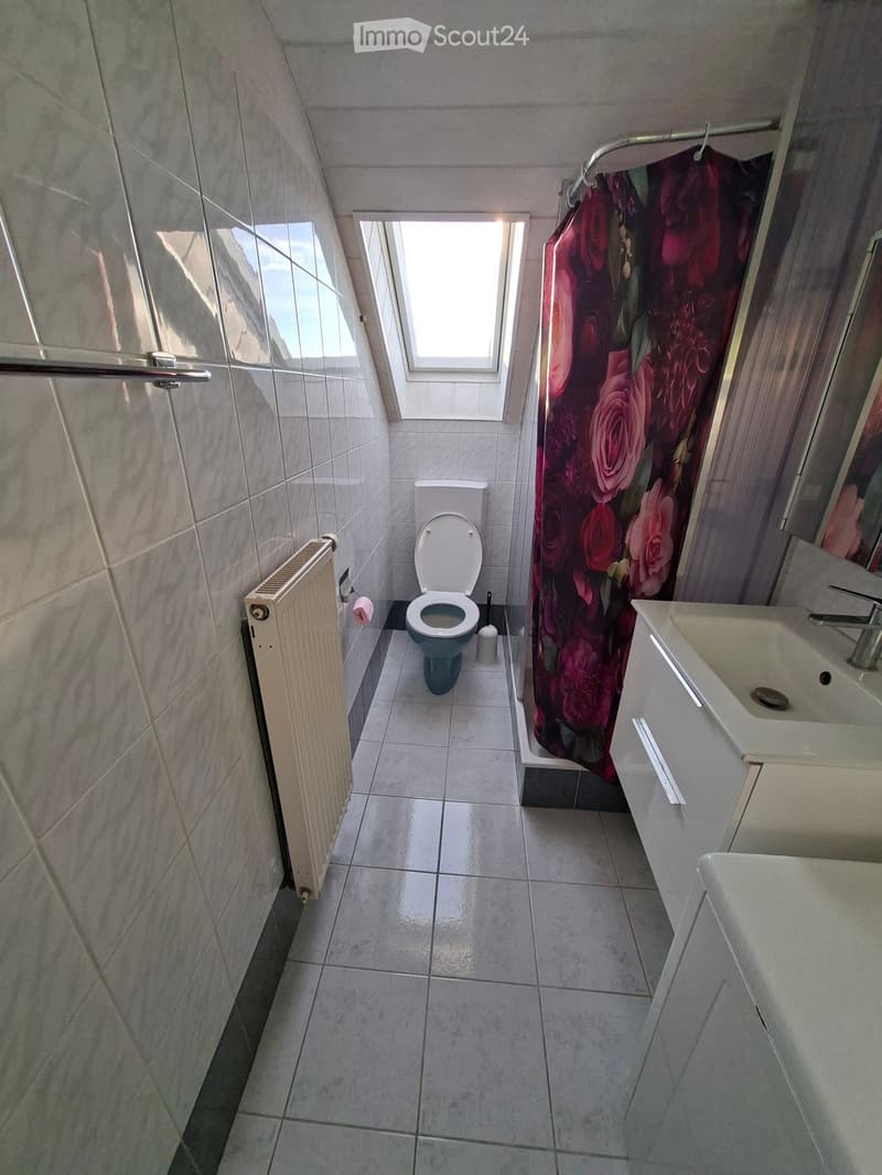 Small bathroom with tiled walls and floor, featuring a toilet, sink, and shower area with a floral curtain. A window provides natural light.