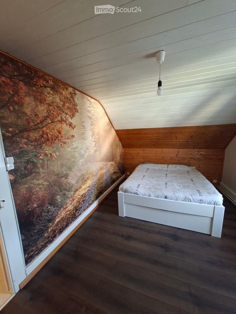 Cozy attic bedroom with wooden slanted ceilings, dark hardwood floors, and a large mural of autumn forest on one wall. A white bed with patterned bedding is positioned against the wooden wall.