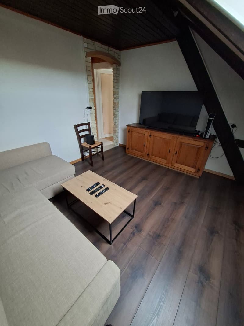 Living room with wooden flooring, a light-colored sofa, a wooden coffee table with remote controls, a wooden TV cabinet, a chair, and a flat-screen TV.