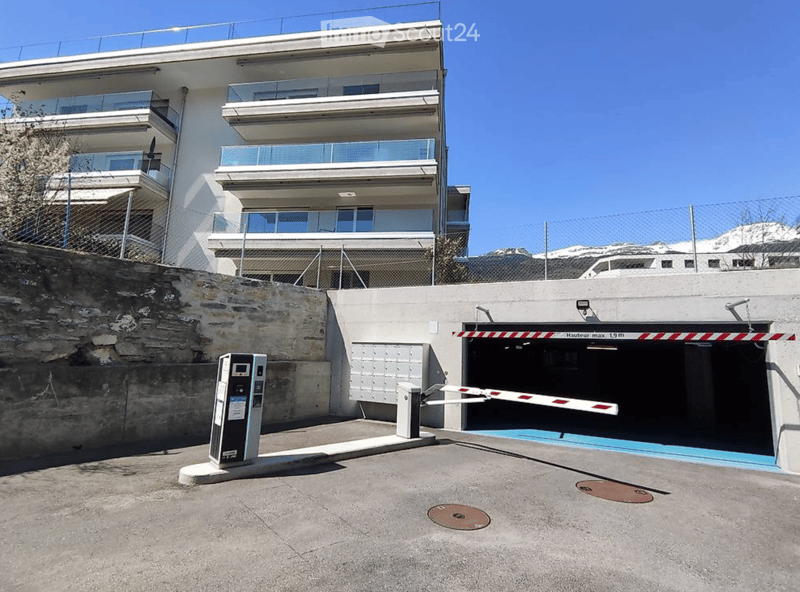 Modern multi-story building with balconies and a gated parking area. The entrance features a security gate and a parking barrier.