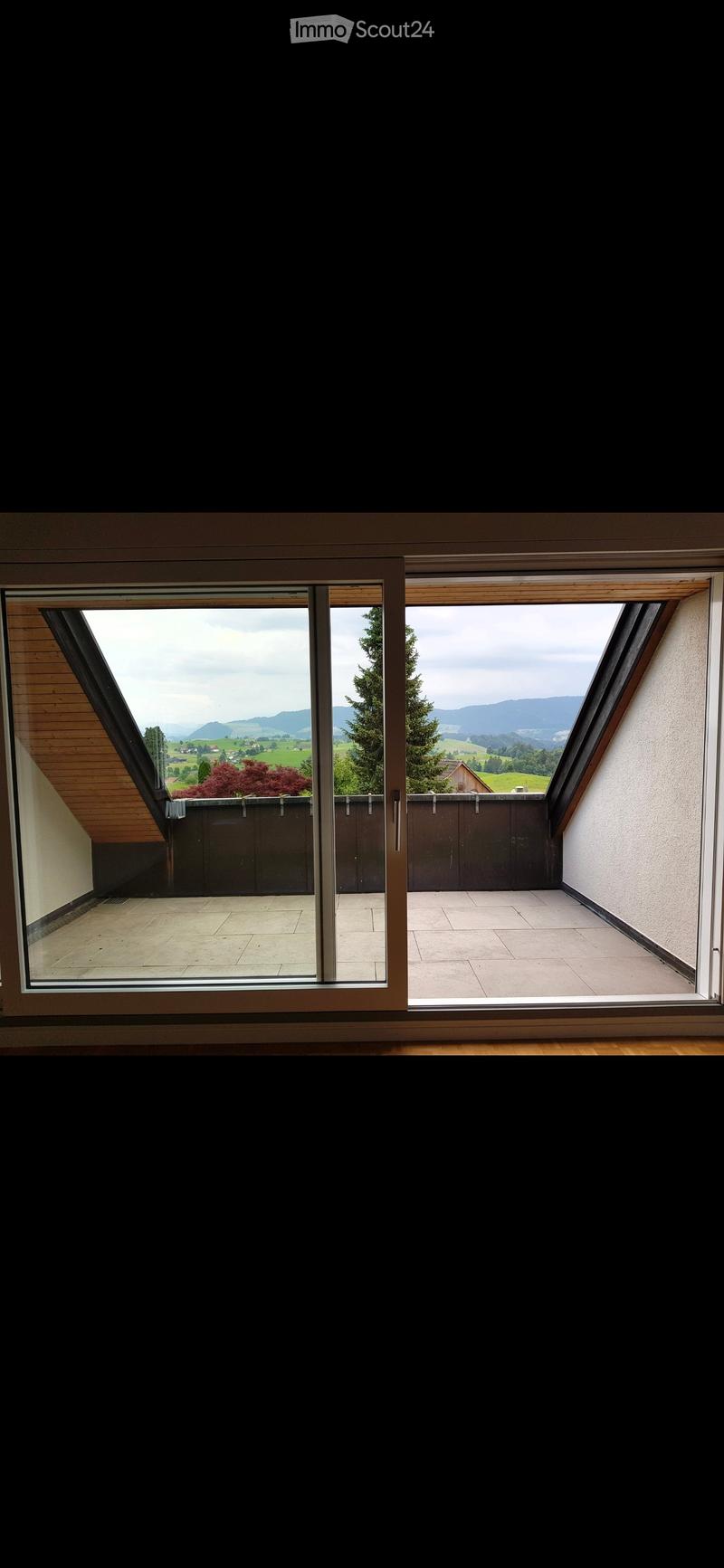 Large sliding glass doors open to a tiled balcony with a view of green fields and mountains.
