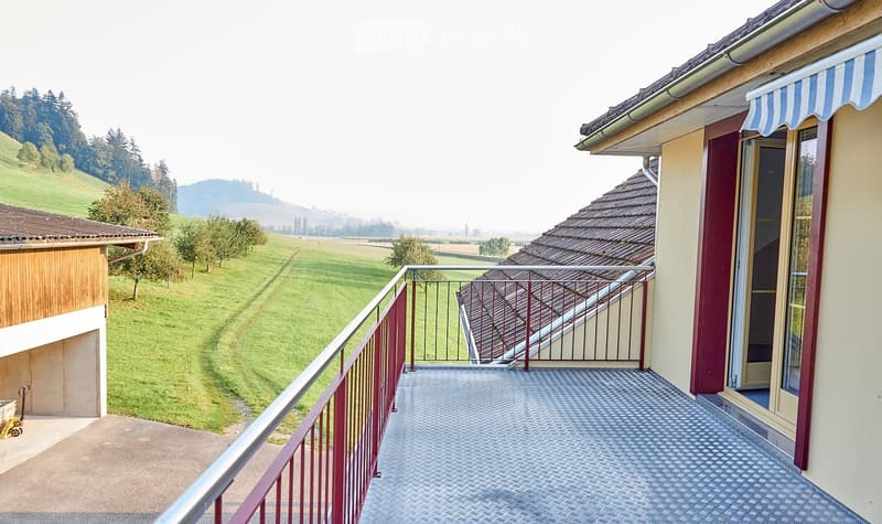 Balcony with metal railing overlooking a green field and distant hills, with a striped awning on the adjacent building.