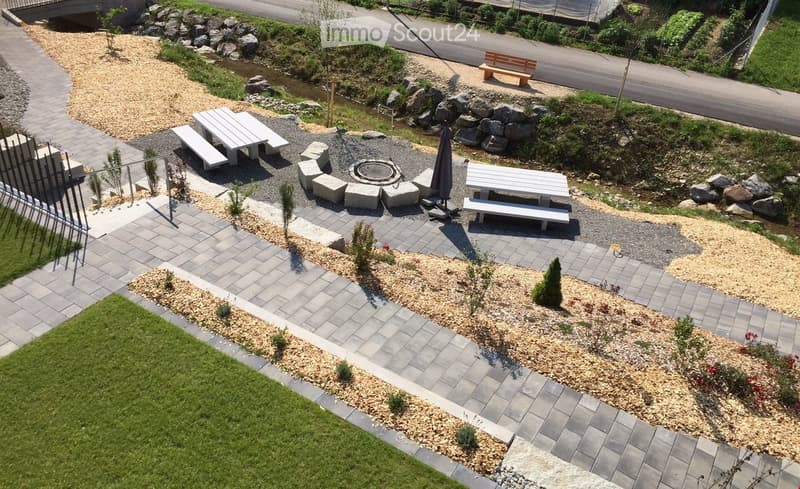 Patio with concrete floor, benches, fire pit, and plants, elevated road, and stream