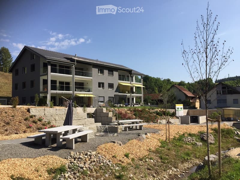 Large grey building, three floors, yellow awnings on first floor, balconies, fenced garden area, picnic tables, cobblestone paths
