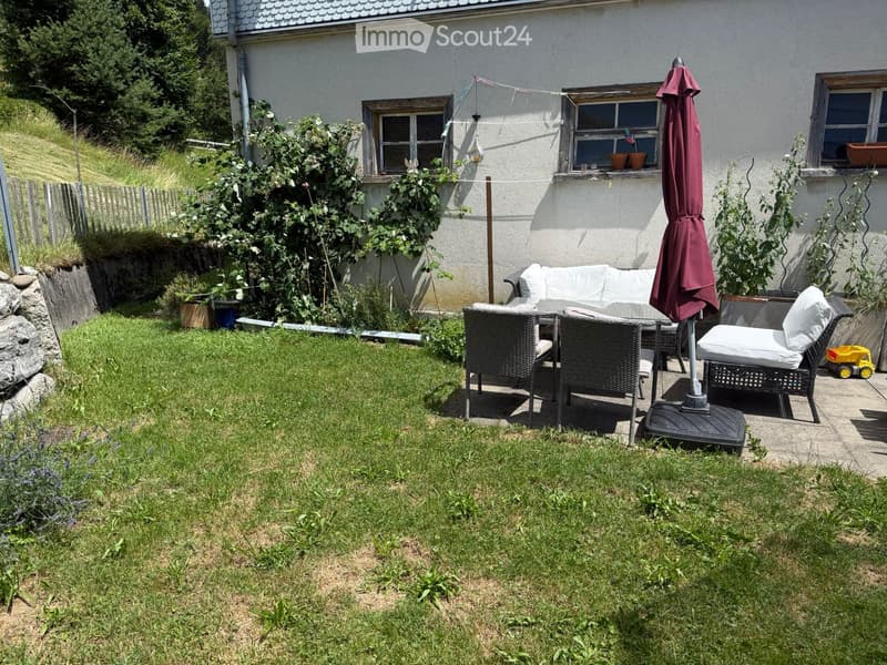 Garden with a grassy lawn, patio seating area with a sofa, chairs, table, and umbrella, and a small toy on the ground.