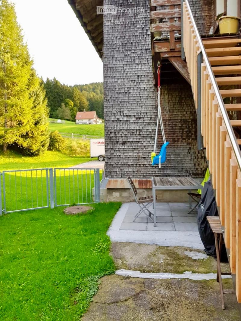 Outdoor area with a wooden table, chairs, and a swing attached to the building. The area is bordered by a metal fence and features a grassy lawn and a gravel pathway.