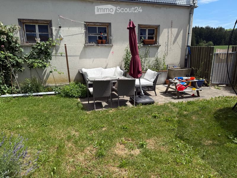 Garden area with a white sofa, outdoor table, chairs, umbrella, and garden tools near the house.