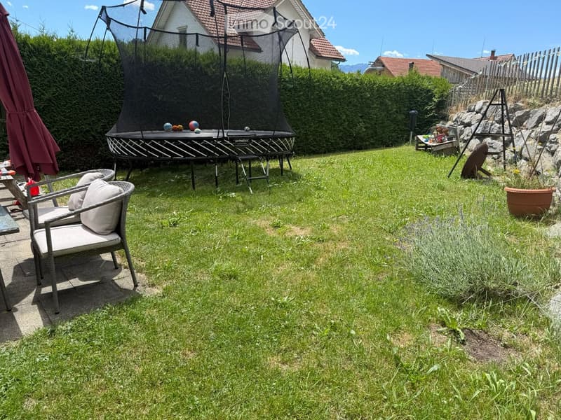 Lush green garden with a trampoline, a wicker chair with cushions, and a large umbrella on a patio.