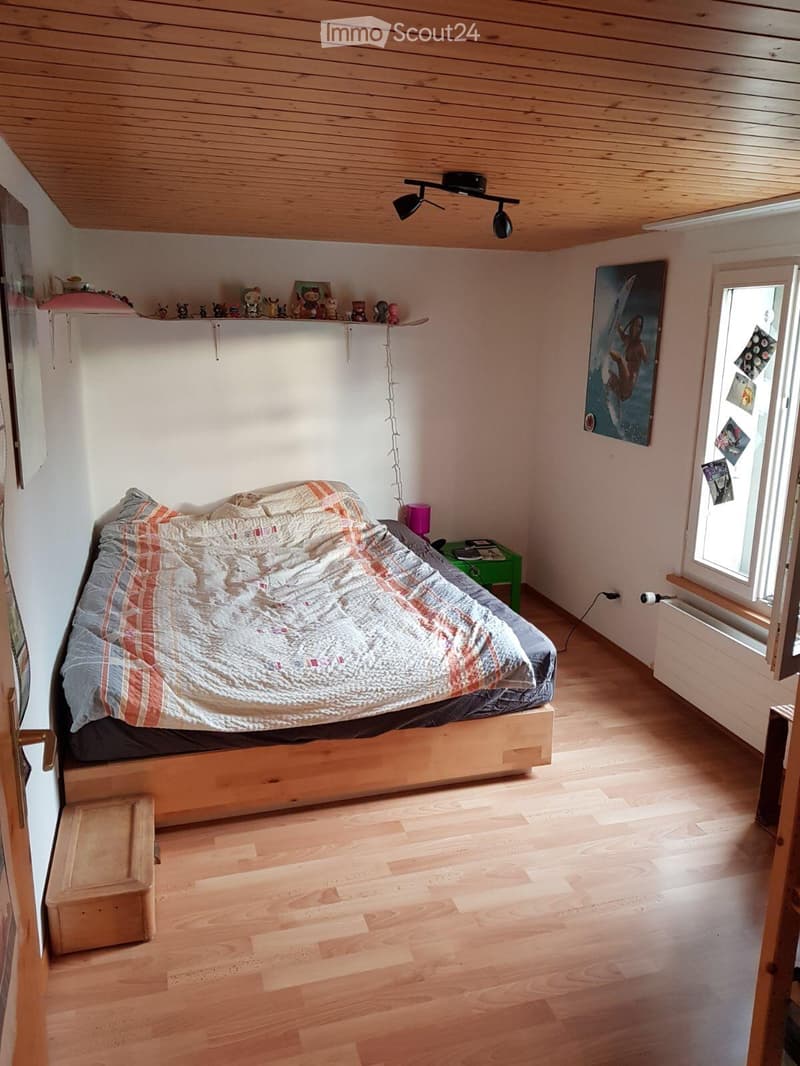 Cozy bedroom with wooden floors, a bed with a colorful quilt, a small green nightstand with a lamp, a shelf with decorative items, and a large window with a view.