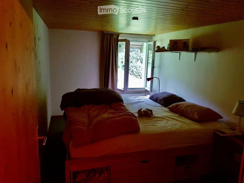 Cozy bedroom with a wooden bed, pillows, a nightstand with a lamp, and a small toy on the bed. The room features wooden walls and a large window with curtains.